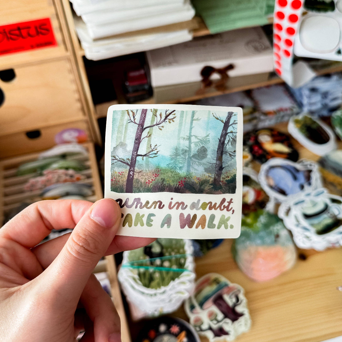Hand holding a sticker with a forest design and text 'When in doubt, take a walk.' against a background of books and stationery items.