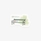 Vinyl sticker of a Copenhagen green bench with a bakery bag and to-go coffee on top and light green background and 'København' as text.