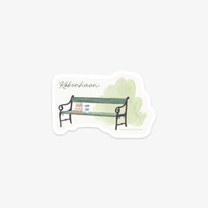 Vinyl sticker of a Copenhagen green bench with a bakery bag and to-go coffee on top and light green background and 'København' as text.